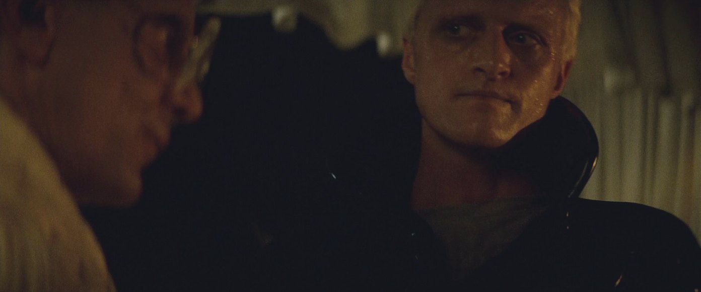 Two men face each other in a dim bedroom lit by candles, the younger one close enough to touch the older one, asking him for something.