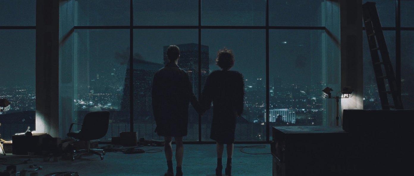 Two people stand in silhouette at a high window at night, holding hands, watching a tower on the skyline beyond the glass begin to fall.