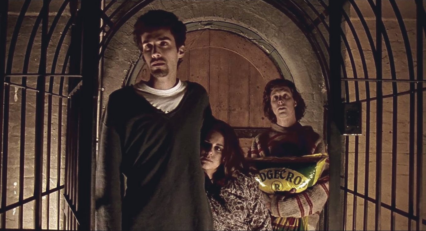 Three people stand in a brick archway behind a barred iron gate at night, the one on the right holding a large sack of garden fertiliser against their chest.