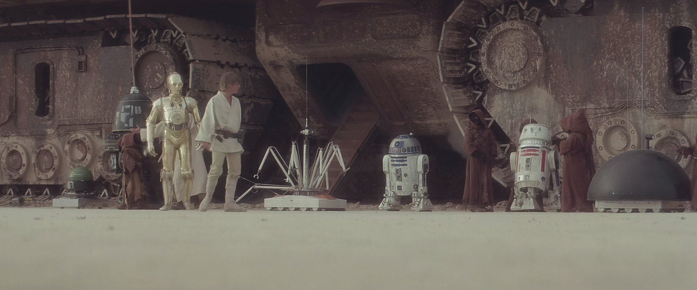 A line of second-hand robots stood out on the sand beside the transport that brought them, while a buyer walks the line and questions the one that talks.