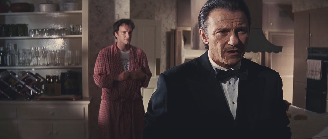 A man in a dinner jacket stands in a suburban kitchen early in the morning, listening, while the owner of the house waits behind him in a dressing gown.