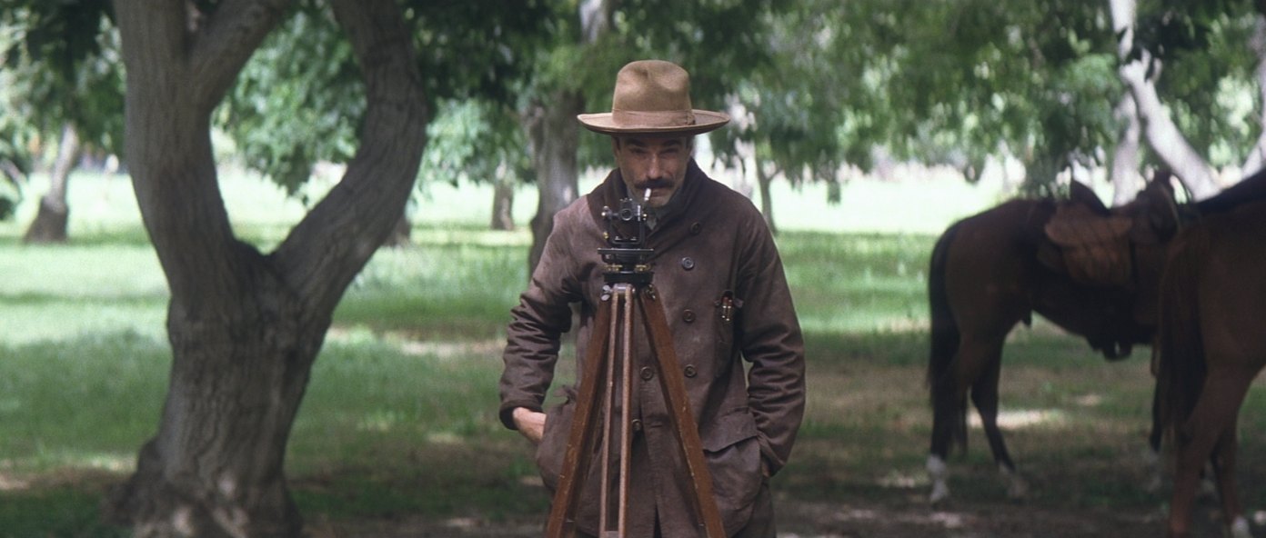 A man in a long coat stands behind a surveyor's theodolite on a wooden tripod in an orchard, with two saddled horses waiting under the trees behind him.