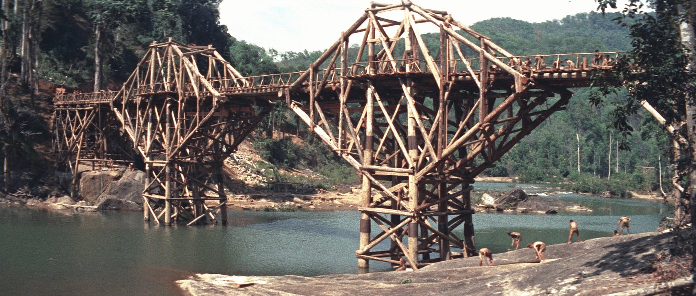 A large timber trestle railway bridge under construction across a jungle river, its deck lined with men at work, with more men on the bare rock in the foreground.